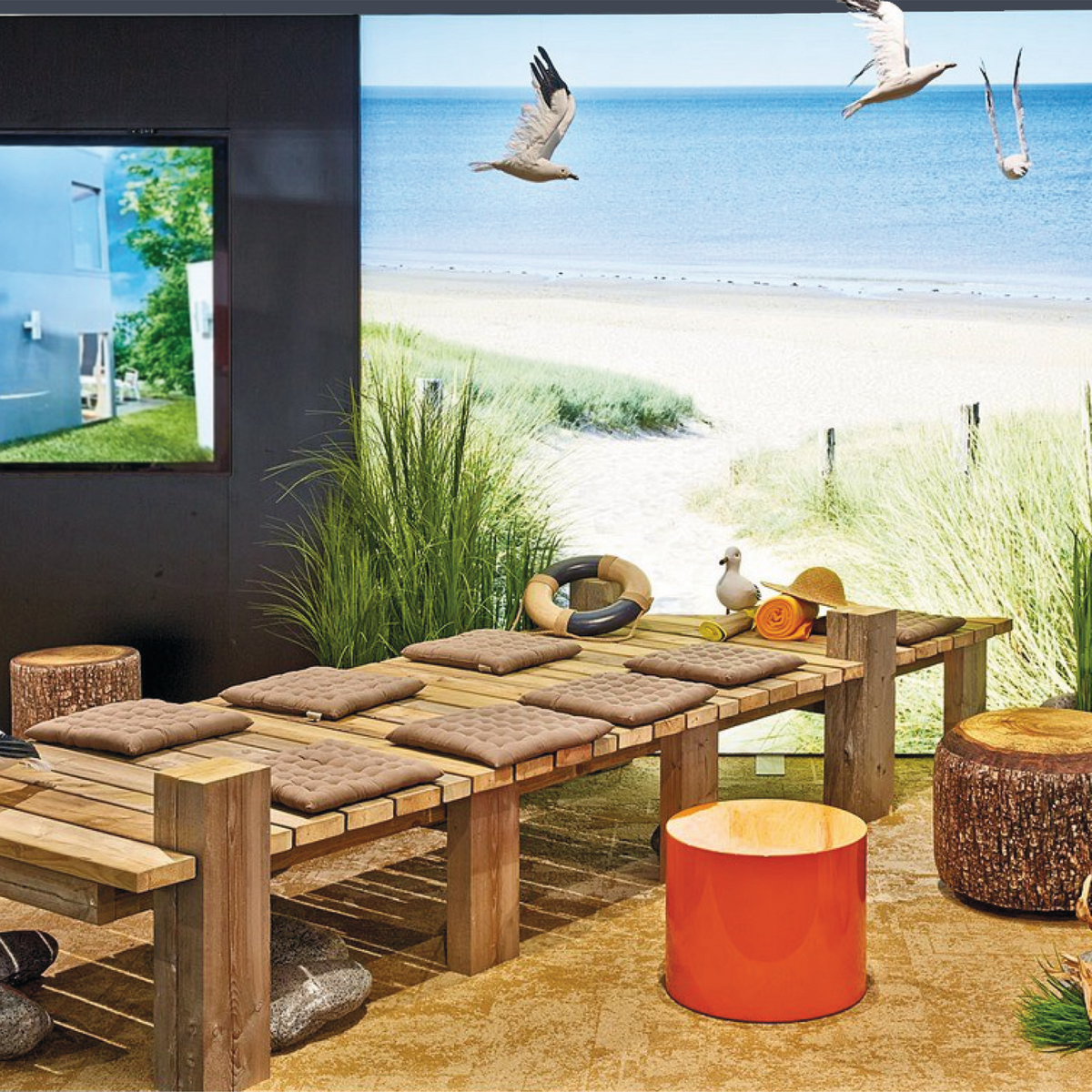 Ein Holzsteg mit Sitzpolstern im Vordergrund, im Hintergrund erstreckt sich der Strand als beleuchtetes Foto